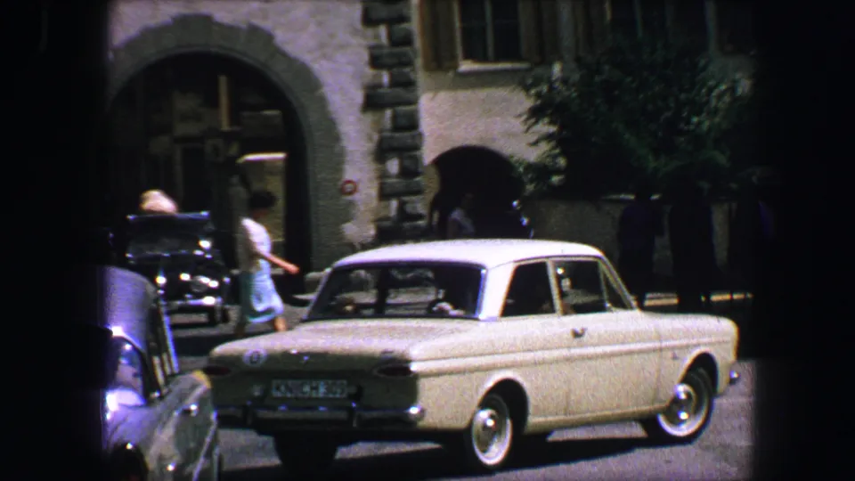 A light-colored mid-century car passes through the Lucerne street scene near a stone arched gate, with pedestrians in the background.