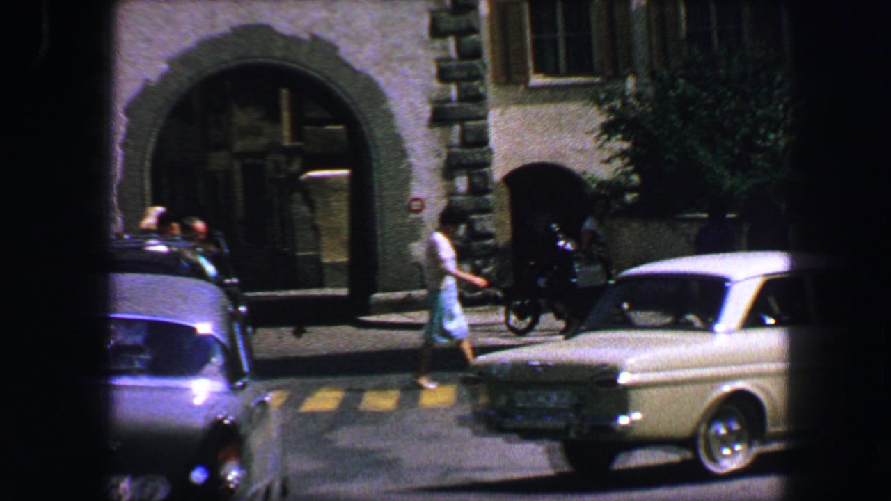 Pedestrians and cars pass a stone building with an arched entrance on a sunny street.