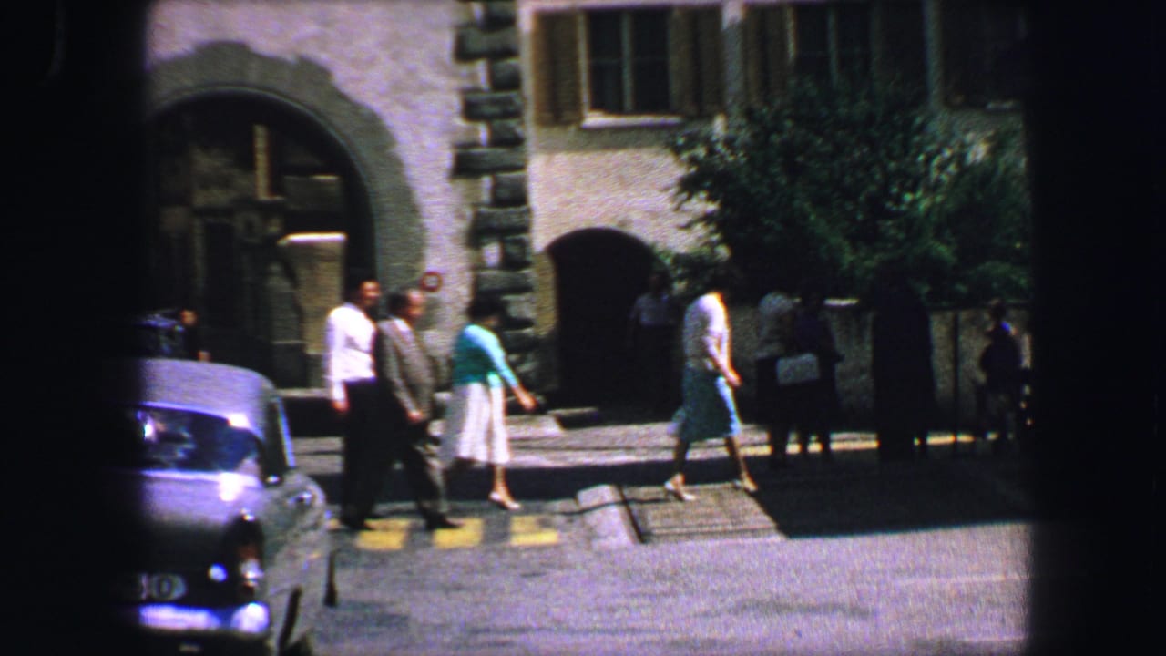 Several adults cross a street in front of an arched stone building and parked cars.