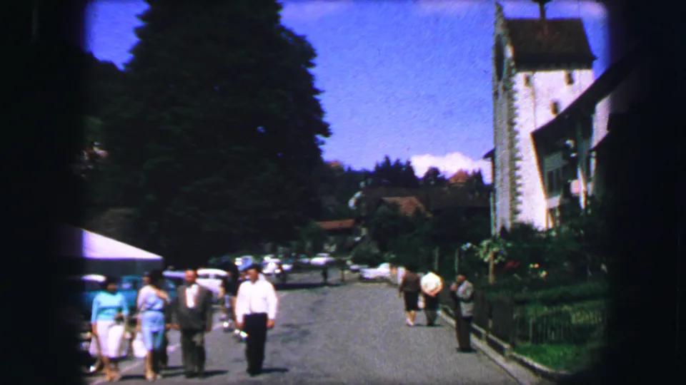 Wider 1963 Lucerne street view with pedestrians near parked cars, trees, and buildings in a sunlit home-movie frame.