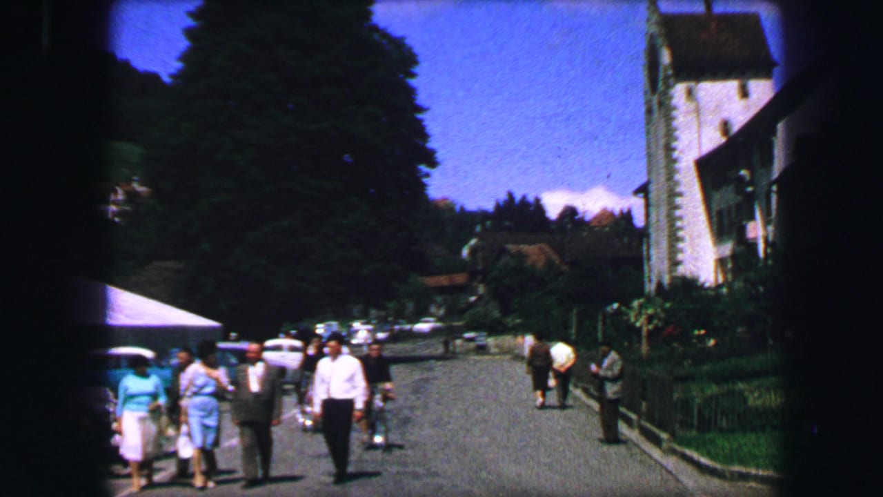 1963 Lucerne street view with pedestrians, parked cars, trees, and a church tower in the background.