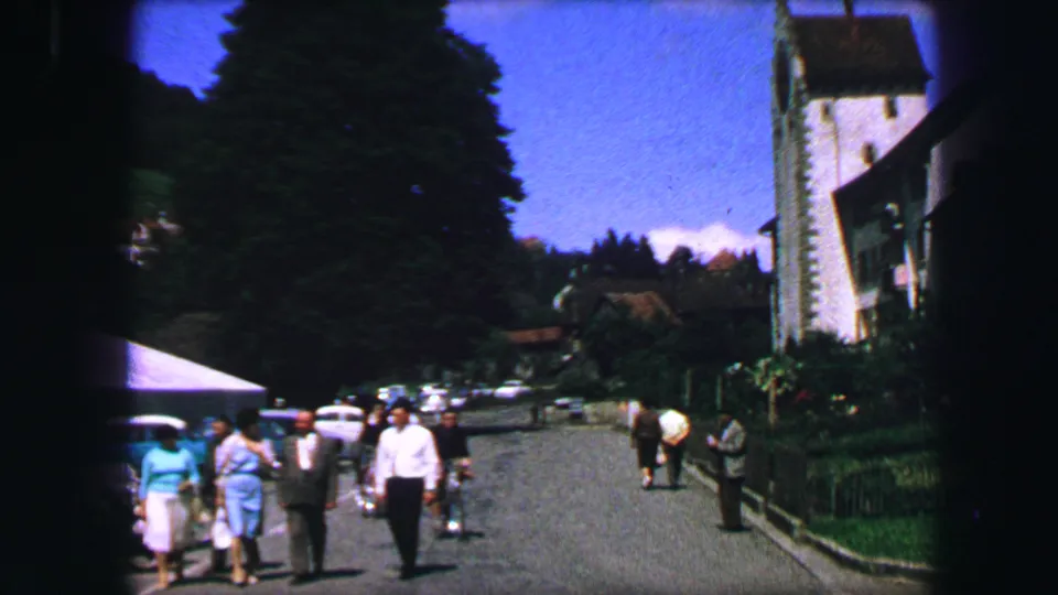 1963 Lucerne street view with pedestrians, parked cars, trees, and a church tower in the background.