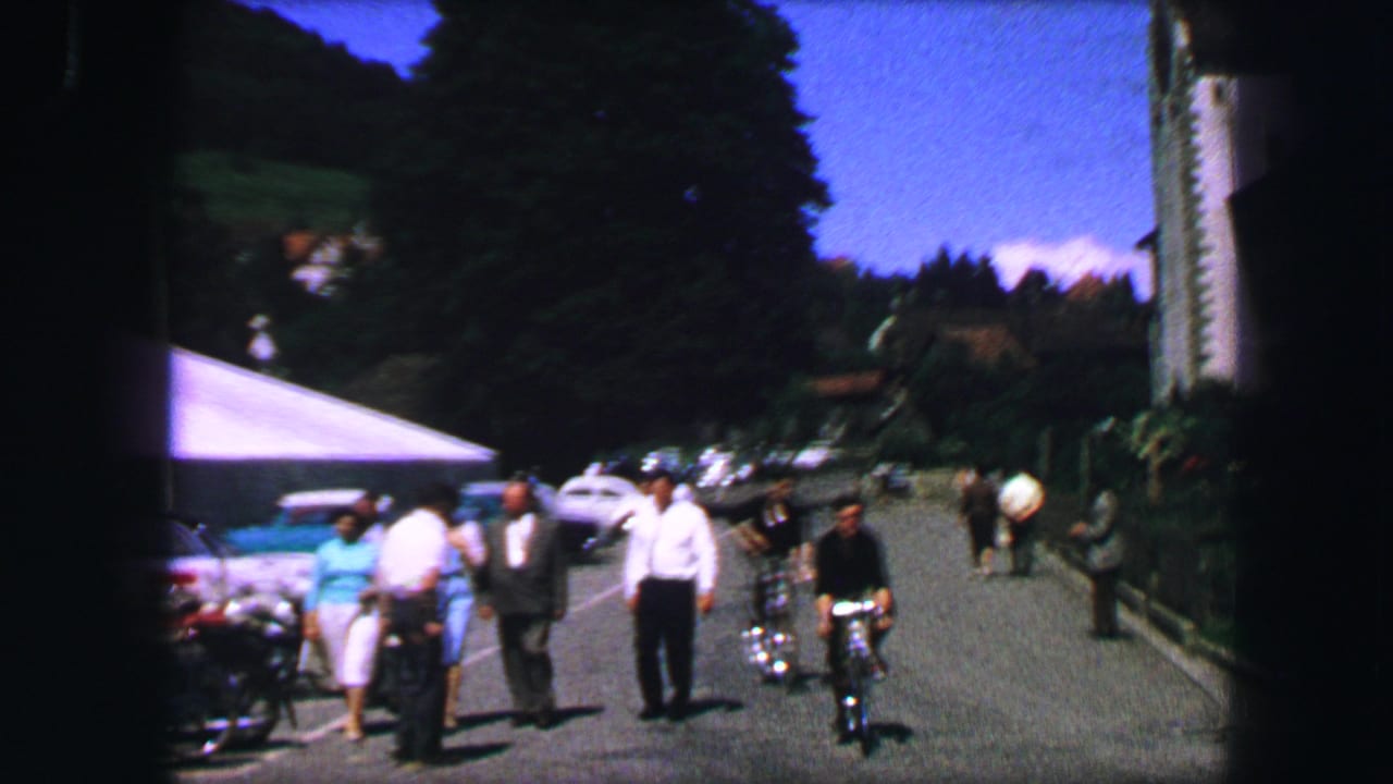 People and a cyclist move along a street beside parked cars and trees.