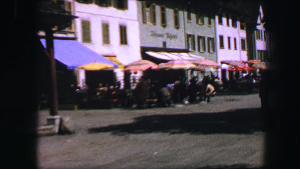Lucerne cafe frontage with colorful umbrellas, seated or standing people, and pale traditional buildings in 1963 home-movie footage.