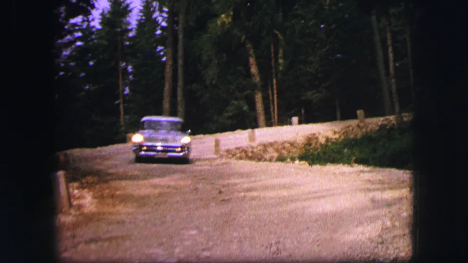 Grey 1960s-era car approaches on a wooded road in archival home-movie footage from Saint Moritz.