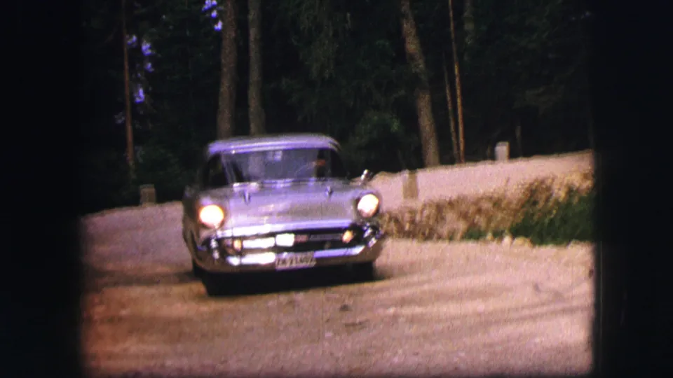 Close front view of a grey mid-century sedan approaching on a forest road in 1963 Saint Moritz footage.