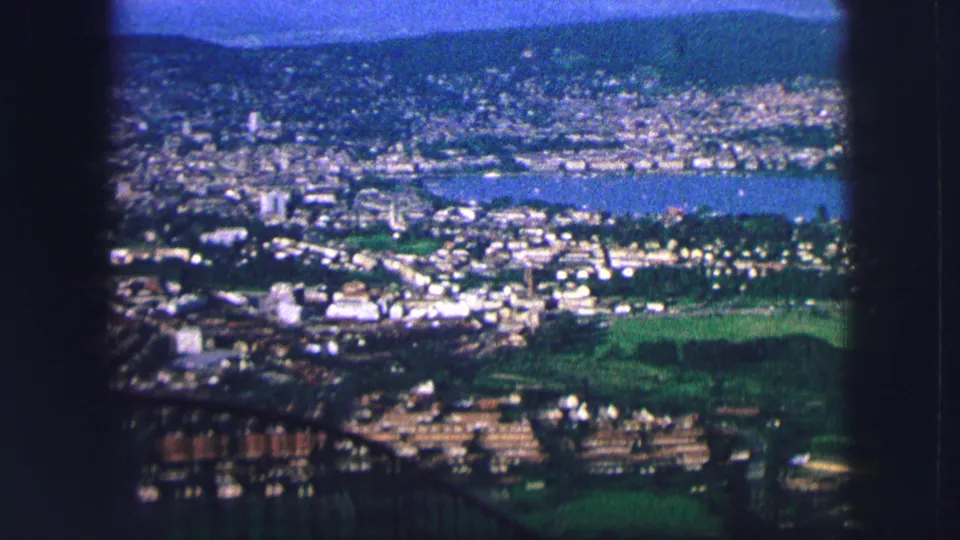Saint Moritz landscape view with the lake, clustered buildings, green foreground slopes, and hills in a grainy 1963 8mm frame.