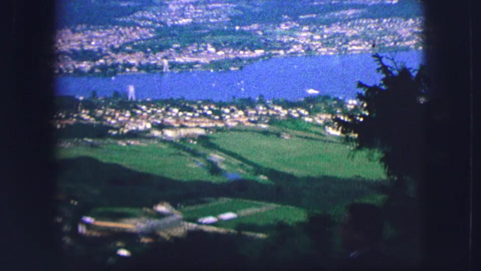 1963 8mm view from Saint Moritz over a blue lake, clustered lakeside buildings, green fields, and shadowed foreground trees.