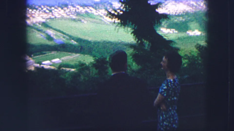 Silhouetted visitors at a Saint Moritz hillside viewpoint look across green fields toward the lake and town in 1963 home-movie footage.