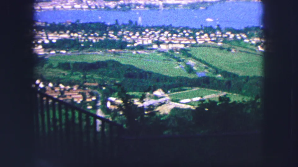 1963 Saint Moritz balcony view with a dark railing in the foreground, green hills and fields below, and the lake behind the town.