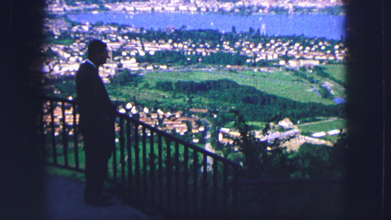 In 1963 Saint Moritz, a suited man stands in silhouette at a high overlook facing a sunlit valley, clustered buildings, and a blue lake.