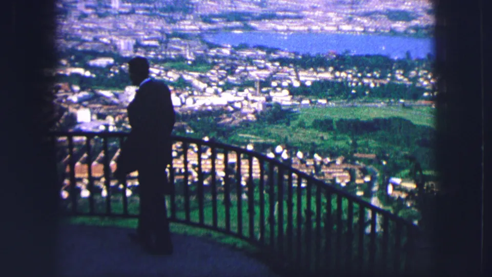 A dark-suited man at the Saint Moritz overlook is framed against the valley settlement, green open land, and blue lake in soft 8mm color.