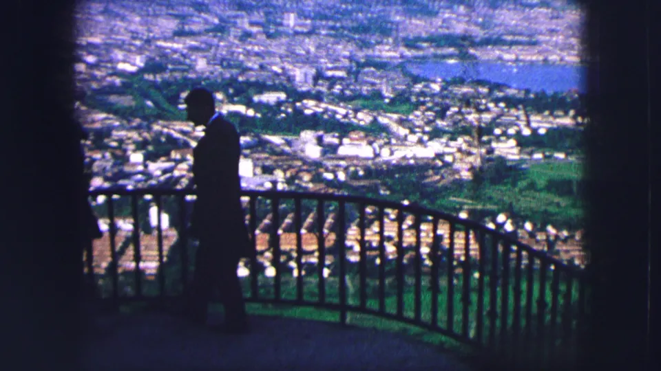 The silhouetted man moves beside the railing at the Saint Moritz viewpoint, with the sunlit valley, buildings, and lake spread behind him.