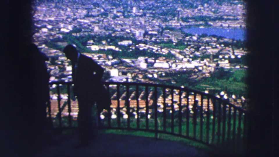 1963 Saint Moritz home-movie frame with a silhouetted visitor at a hillside overlook above town, water, and greenery.