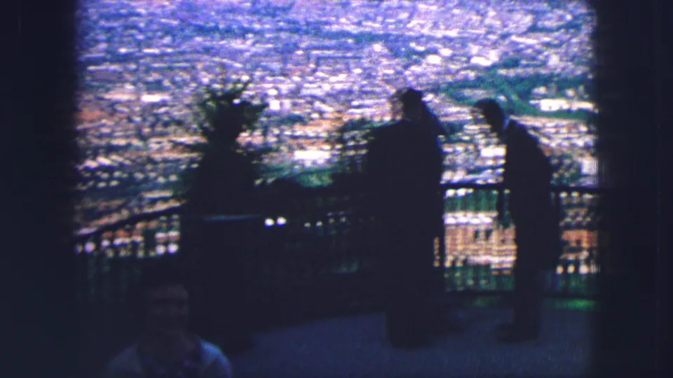 1963 Saint Moritz overlook view with silhouetted visitors near a railing and the cityscape visible in bright soft focus behind them.