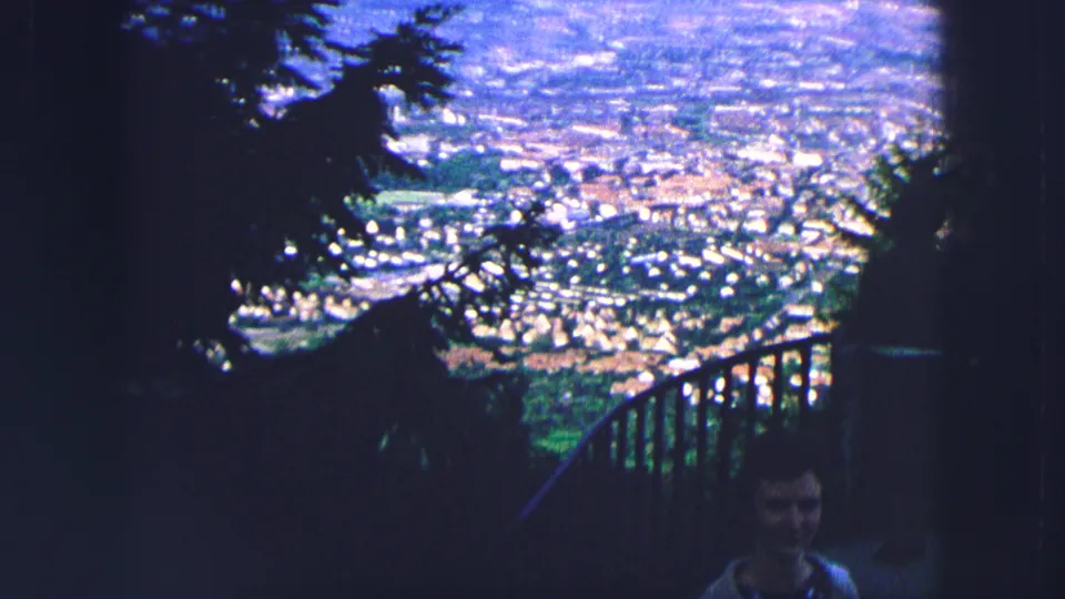 1963 Saint Moritz hilltop view framed by trees and railing, with a partially visible person below the distant townscape.
