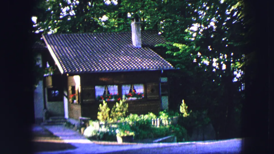1963 Saint Moritz home-movie frame showing a chalet-like hillside house, garden plantings, surrounding trees, and a paved road.