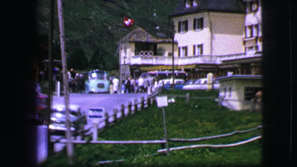 Daylight Swiss resort view with a white hotel building, blue coach, parked cars, fenced lawn, and wooded mountain backdrop.