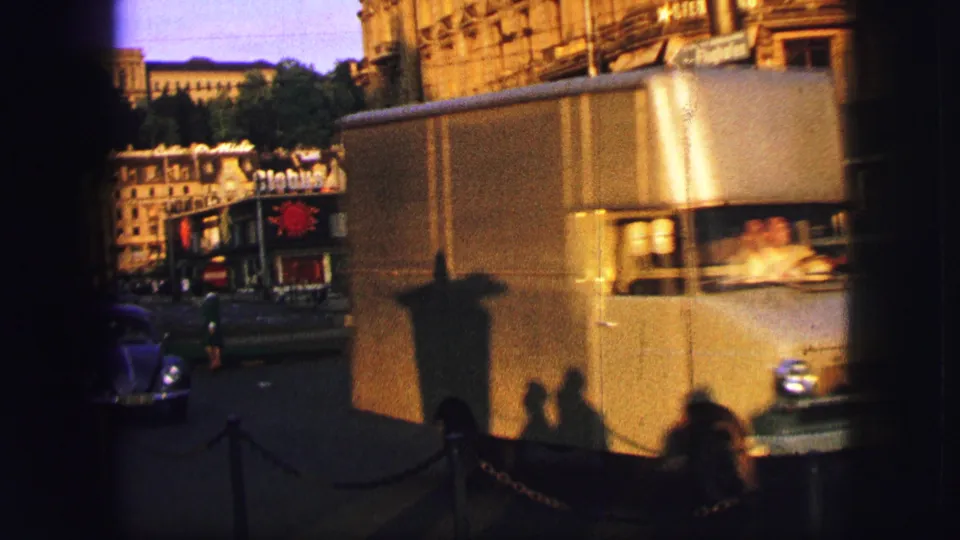 Zurich street scene from 1963 with public transport, compact cars, storefronts, and strong late-day shadows across the road.