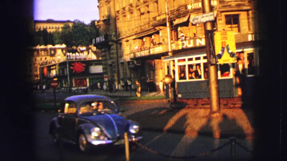 1963 Zurich street view with a compact car in the foreground, trams, shop signs, and distant pedestrians near an intersection.