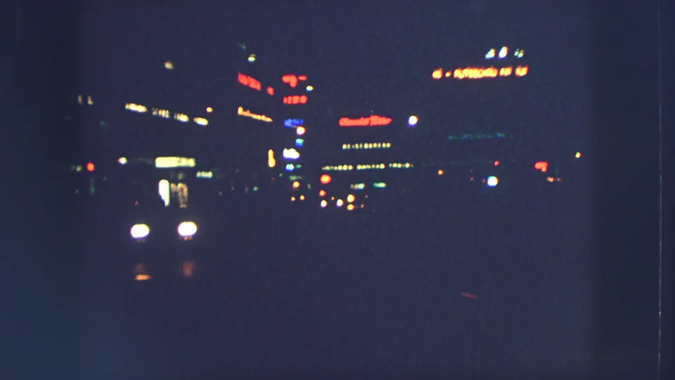 Nighttime Zurich street frame from 1963 showing vehicle headlights, blurred traffic, and illuminated commercial signs in the distance.