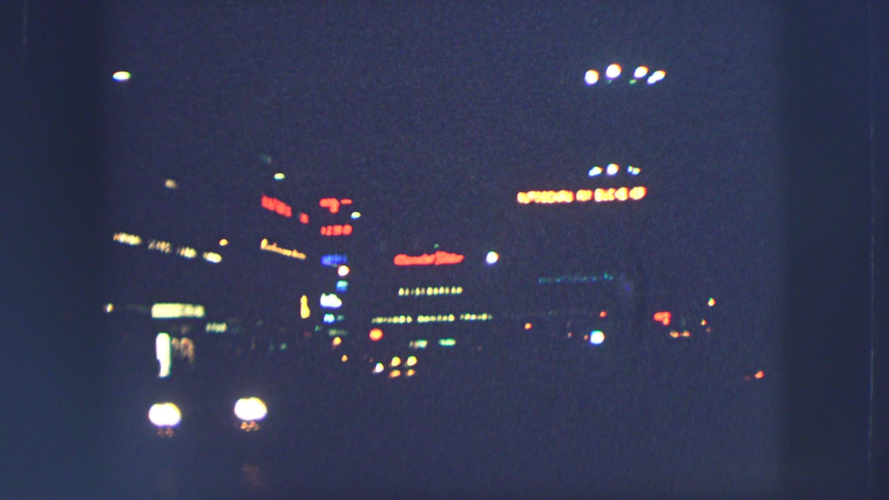 1963 Zurich night street view with soft car headlights, illuminated signs, and deep blue shadows on 8mm film.