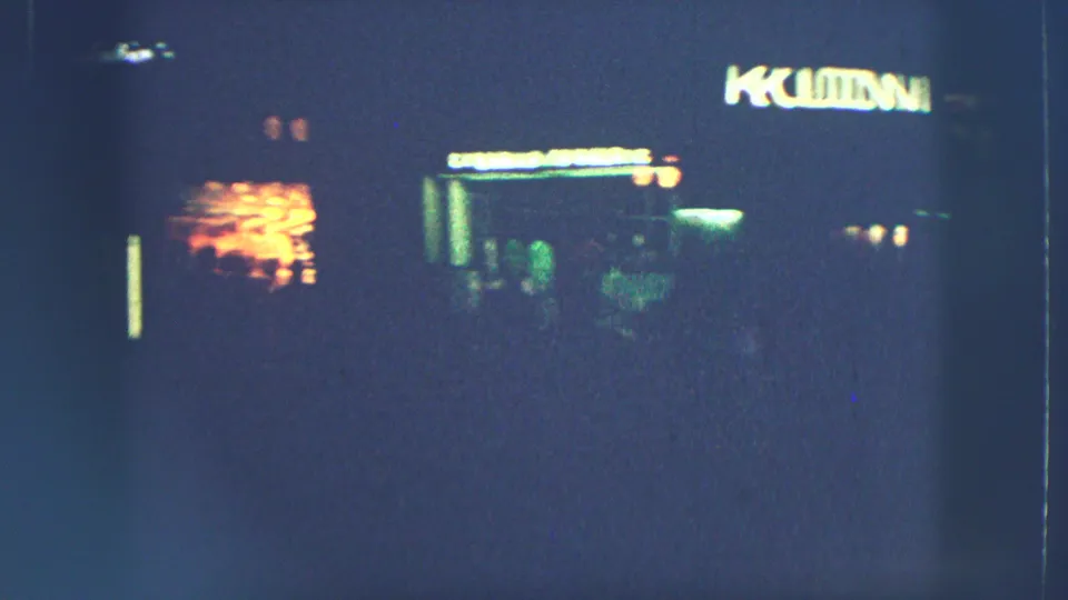 Zurich nighttime storefronts appear in soft focus, with a bright sign, dark pavement, and scattered lights on 8mm film.