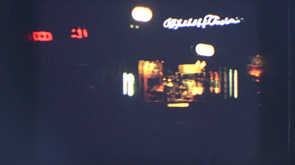 1963 Zurich night view with glowing shopfront lights, blurred signage, and heavy shadow in a handheld 8mm frame.