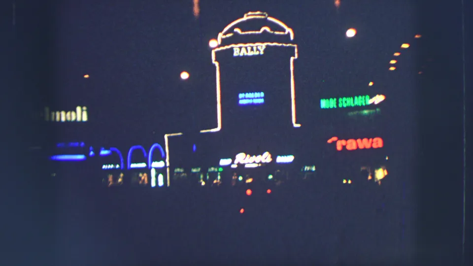1963 Zurich street facade with a bright Bally sign, nearby neon lettering, and a dark roadway in the foreground.