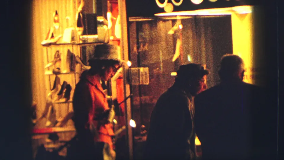 Zurich night storefront in 1963 with warm display lighting and indistinct pedestrian silhouettes moving past the window.