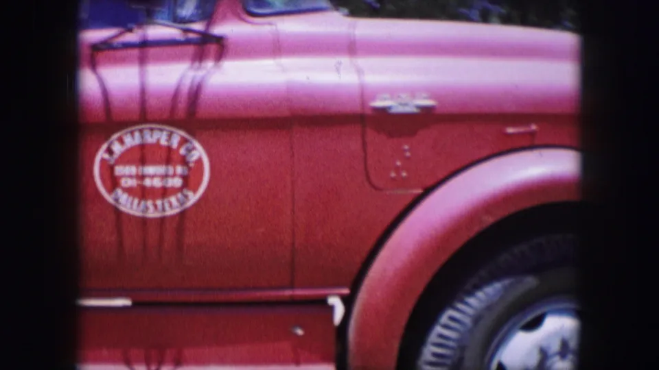 1964 Dallas 8mm frame of the red truck’s front side, including oval painted lettering, fender, wheel, door seam, and a dark frame bar.
