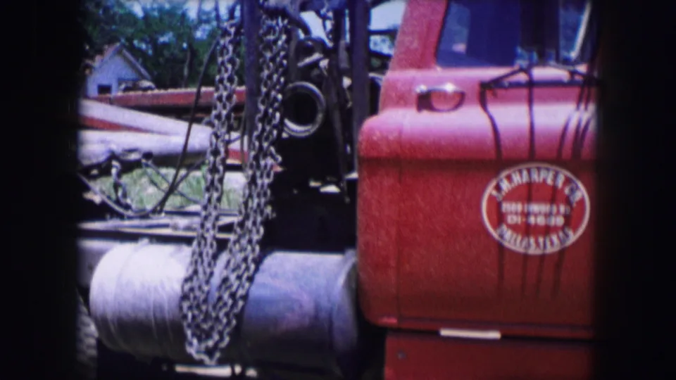 Close 8mm view of chains, rear equipment, and the red truck cab door with a circular logo in a 1964 Dallas scene.