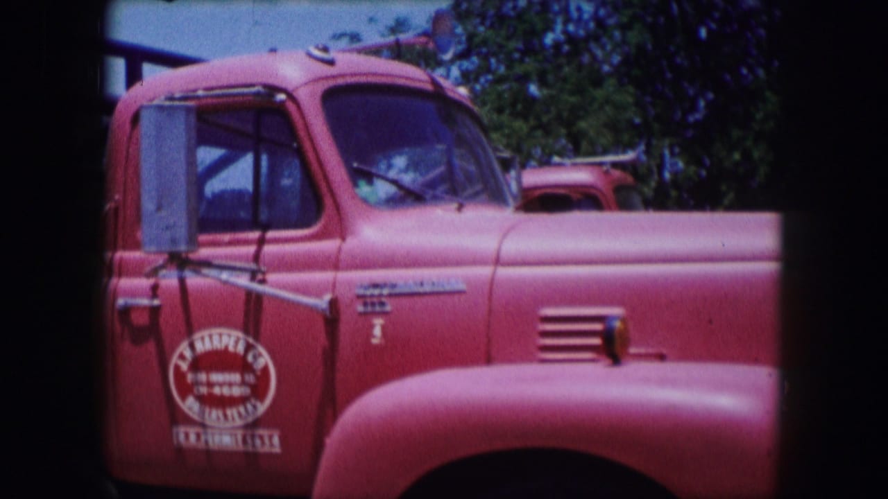 Red truck cab and hood with a door logo, side mirror, windshield, and trees behind it.