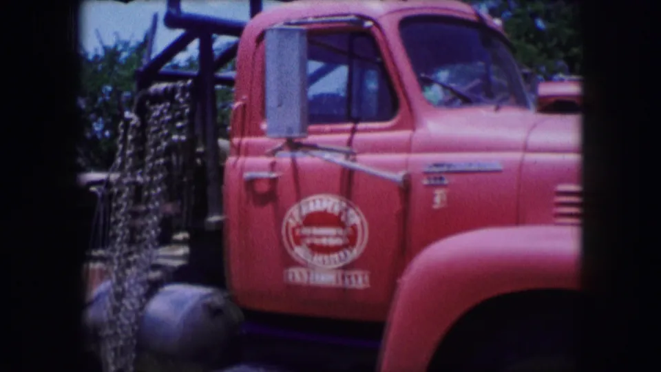 1964 Dallas 8mm frame showing the red industrial truck near dark structural bars, with its door logo, mirror, cab window, and front fender visible.