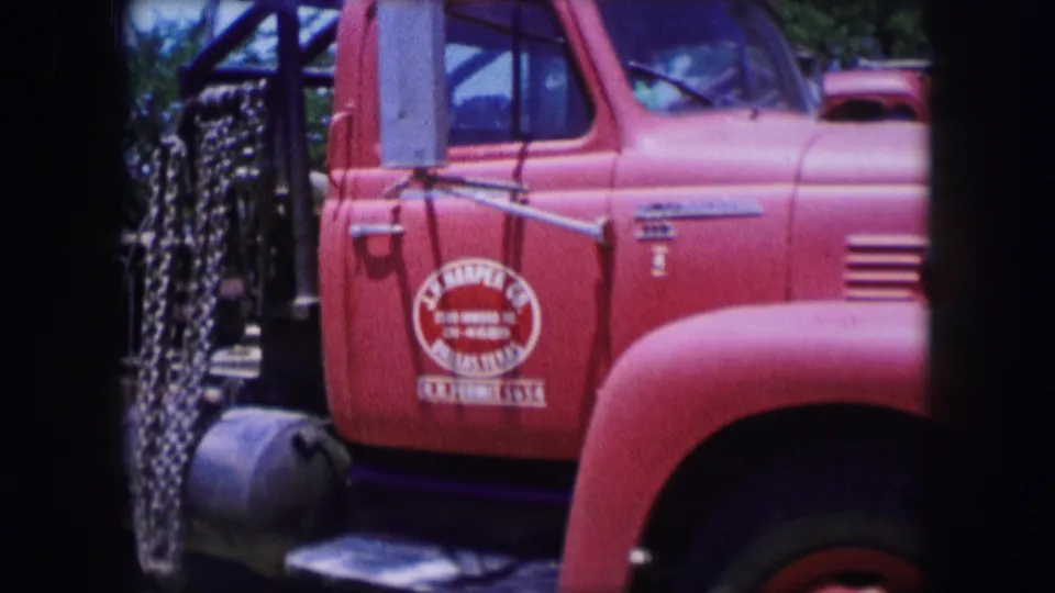 1964 Dallas 8mm frame showing the red truck cab and door lettering, with dark frame masking and industrial equipment partly visible behind.