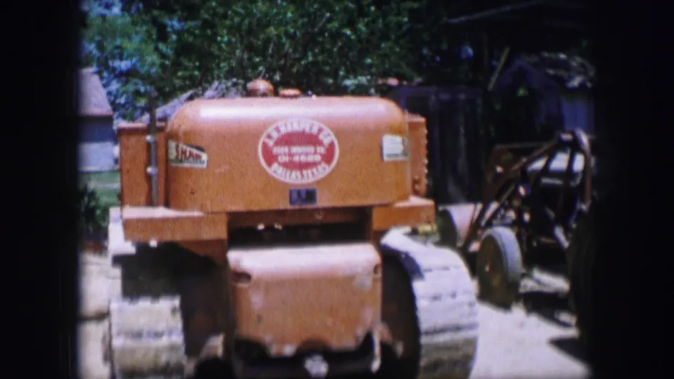 1964 Dallas 8mm frame showing a yellow tracked industrial machine outdoors, with rear lettering, metal treads, trees, and worksite objects nearby.