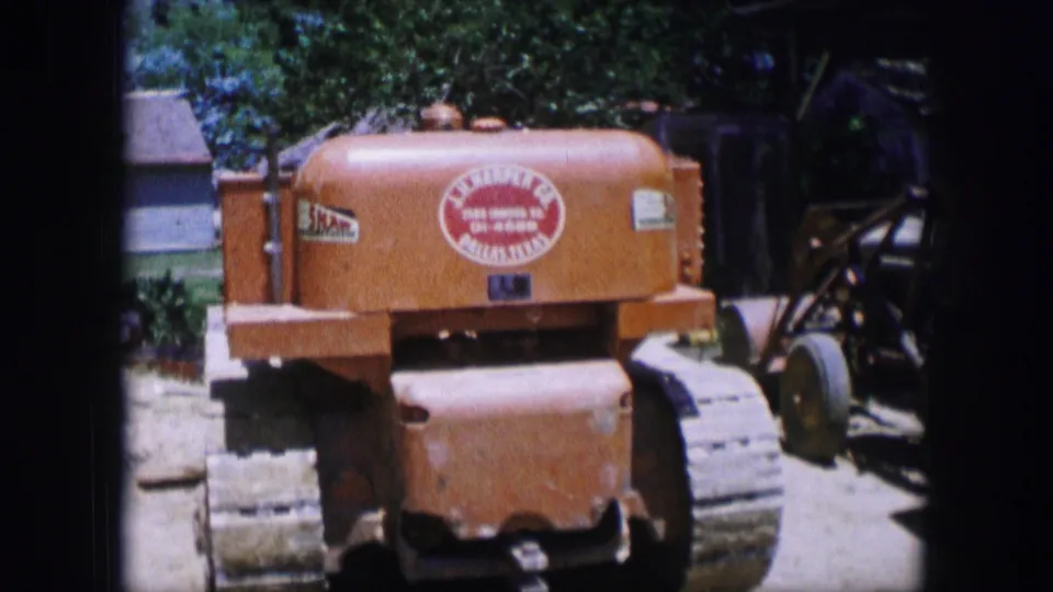 1964 Dallas 8mm frame showing an orange tracked tractor-like machine parked in a sunny yard, with a red circular logo visible.