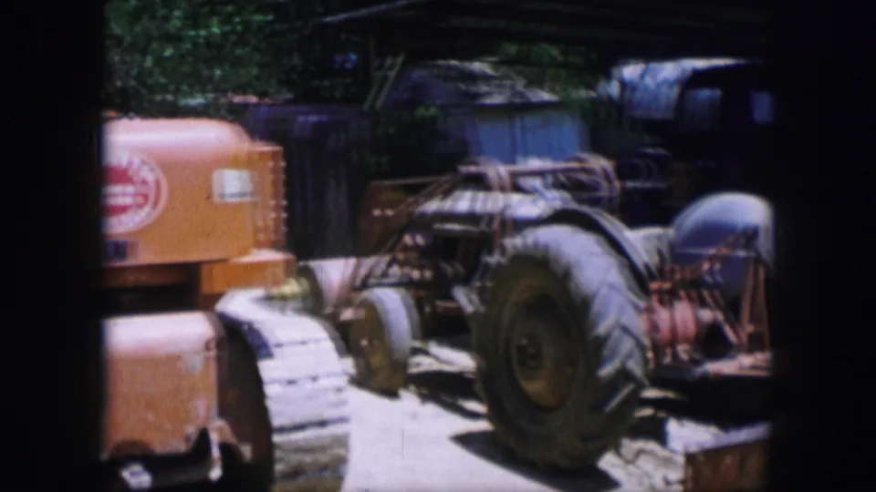 1964 Dallas home-movie frame with the orange tracked machine partly visible beside large rubber tires and parked yard equipment.