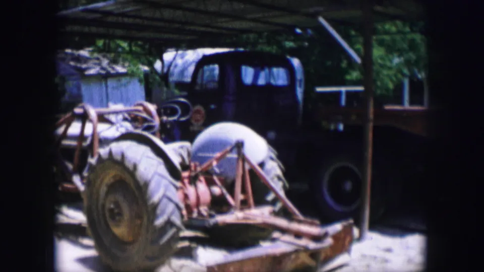 1964 Dallas 8mm frame showing a large tractor wheel, exposed machinery, and a dark older truck parked under a simple shed roof.
