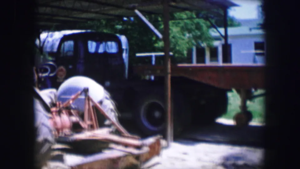 1964 Dallas home-movie frame with a large tractor wheel, exposed machinery, a dark truck, and a flatbed trailer near a shed.