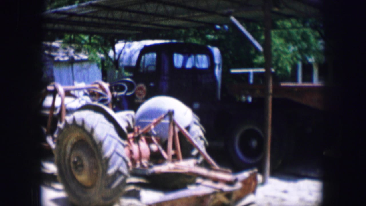 Tractor parked under a shelter beside a flatbed trailer and trees.