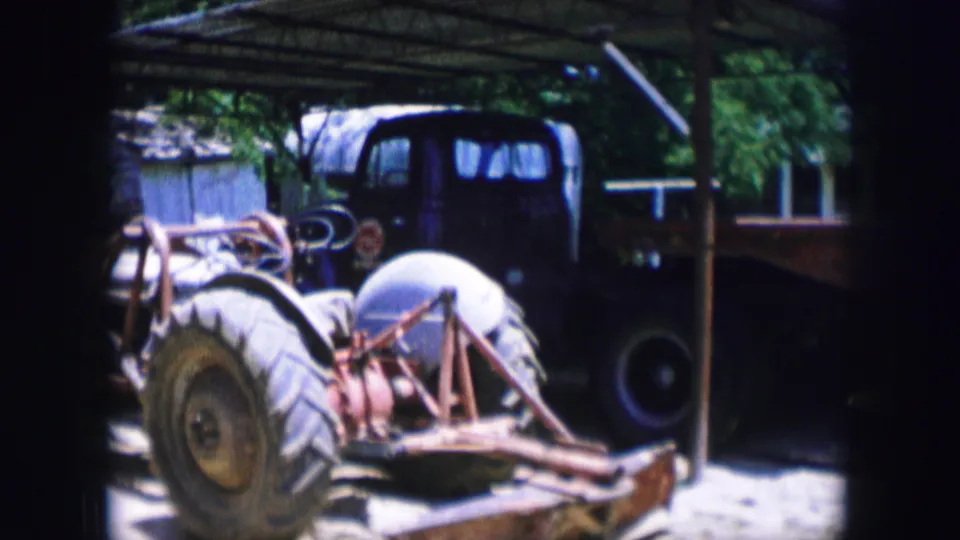 In Dallas, Texas, 1964 home-movie footage, a tractor sits under a simple shelter near a red flatbed trailer and shaded yard.