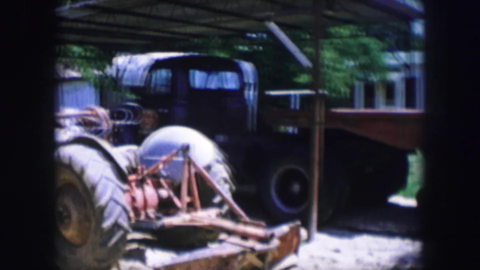 The tractor remains parked near the flatbed trailer, with trees, a shelter roof, and a modest building visible in the background.
