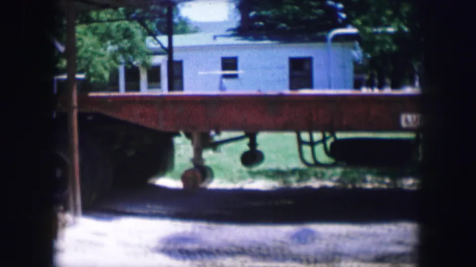 A red flatbed trailer fills the view, with a white building, grass, trees, and part of the tractor edge visible behind it.