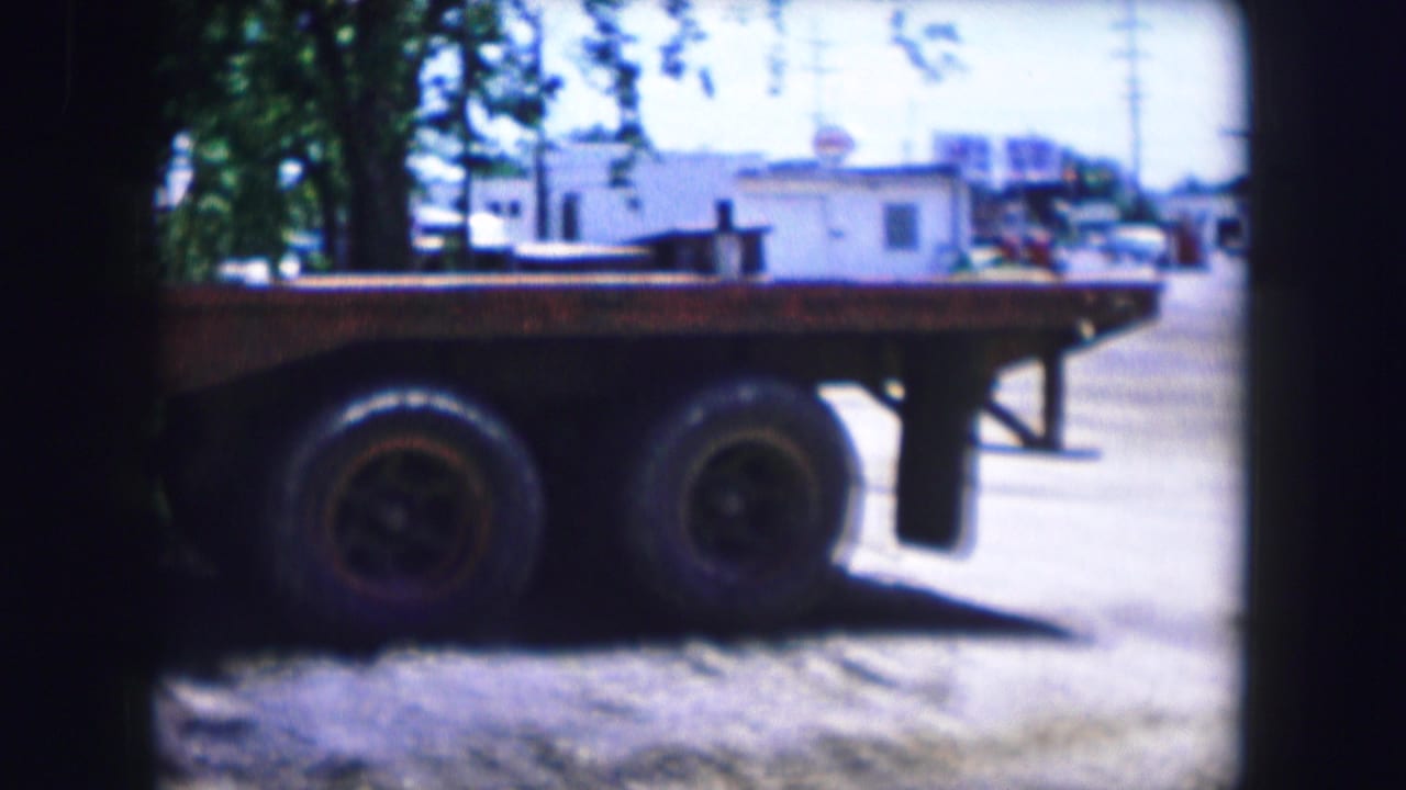 Dallas, Texas 1964 8mm frame showing a large flatbed trailer stationary on a dirt lot near trees and low buildings.