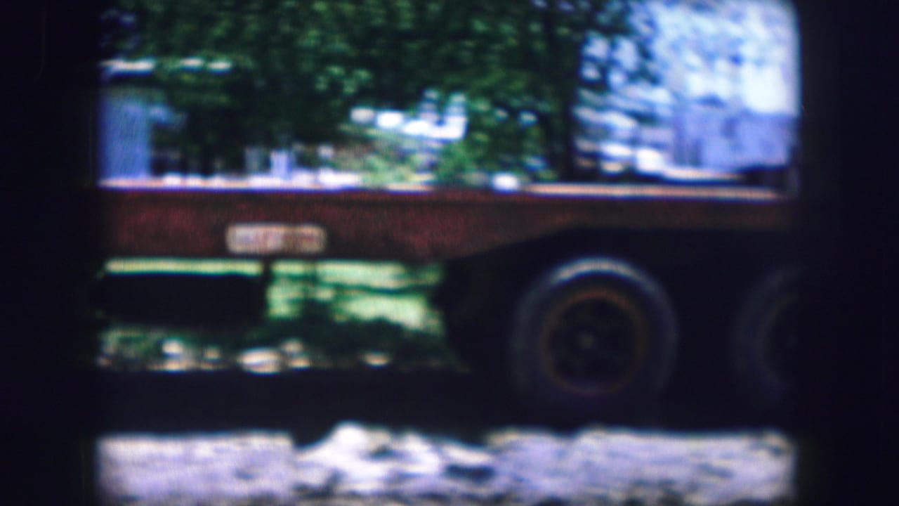 Side of a flatbed trailer partly shaded by trees, with dirt ground below.