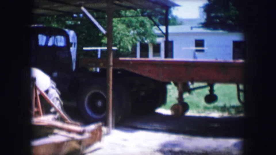 Dallas, Texas 1964 8mm frame with an older dark truck cab, flatbed trailer, dirt lot, trees, and a pale building behind.