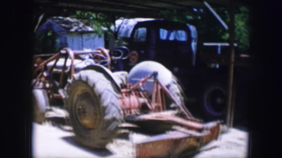 Dallas, Texas 1964 frame showing an older truck cab, exposed machinery parts, large tires, and a flatbed trailer in a shaded yard.