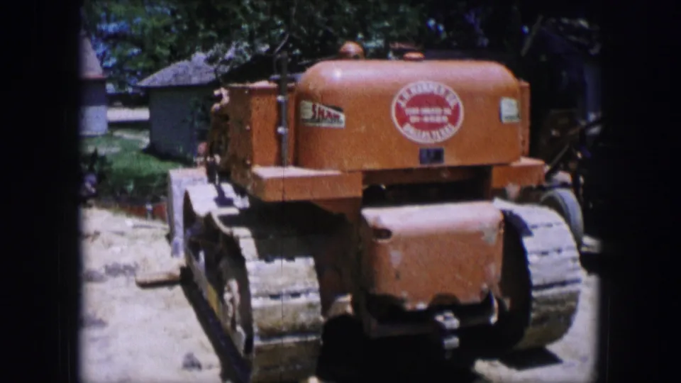 1964 Dallas 8mm frame of an orange crawler tractor parked on dirt, with visible tracks, decals, trees, and a small building in the background.