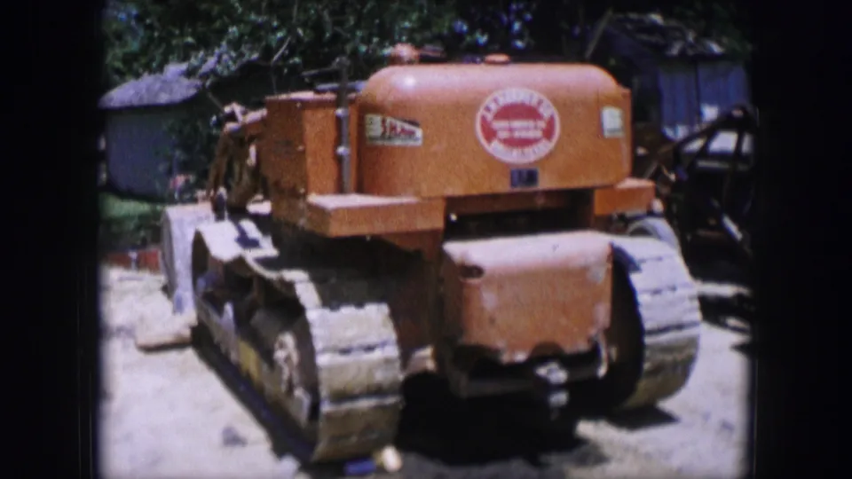 1964 Dallas frame showing an orange tracked tractor from the rear beside other equipment, with sunlit dirt, foliage, and small structures nearby.